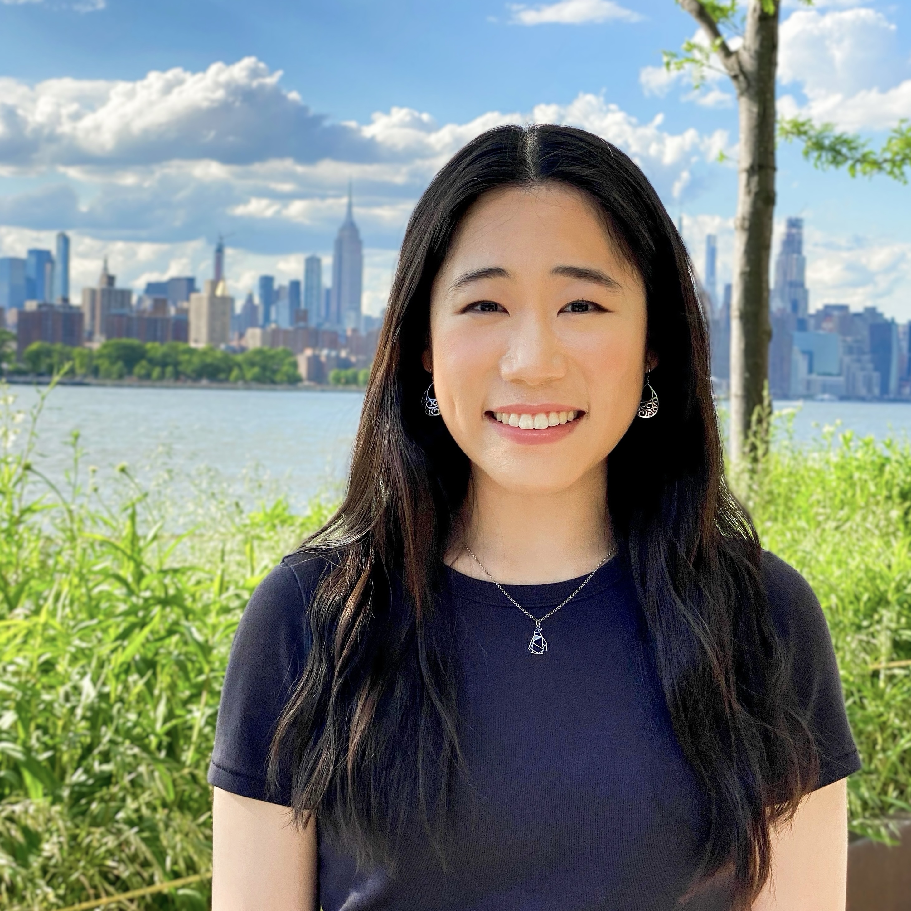 Headshot of Lucy, a Chinese American woman wearing a gray shirt, with the Manhattan skyline and light green foliage in the background.