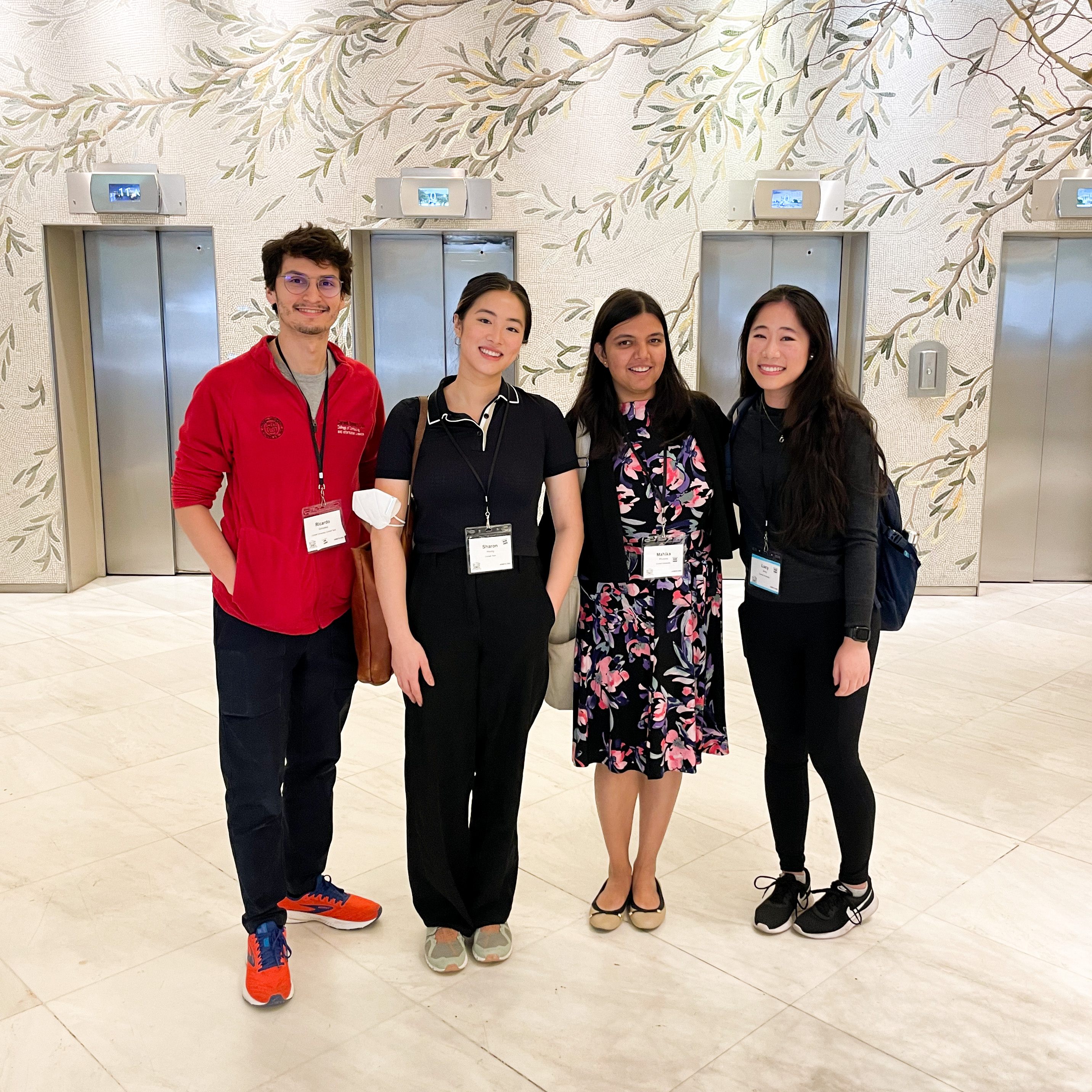 Ricardo, Sharon, Mahika, and Lucy smile and stand together, with an olive branch mosaic and elevators in the background.