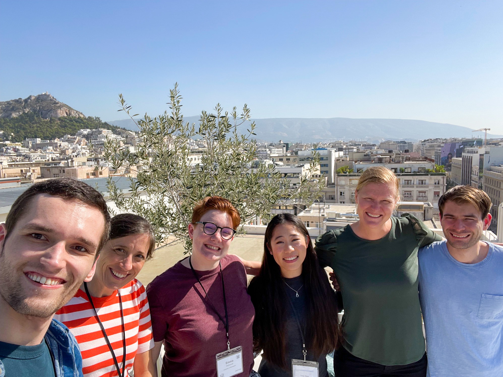 A selfie of Steven, Leah, Emma, Lucy, Abigale, and Adrian, with grey buildings and a tall hill in the background.