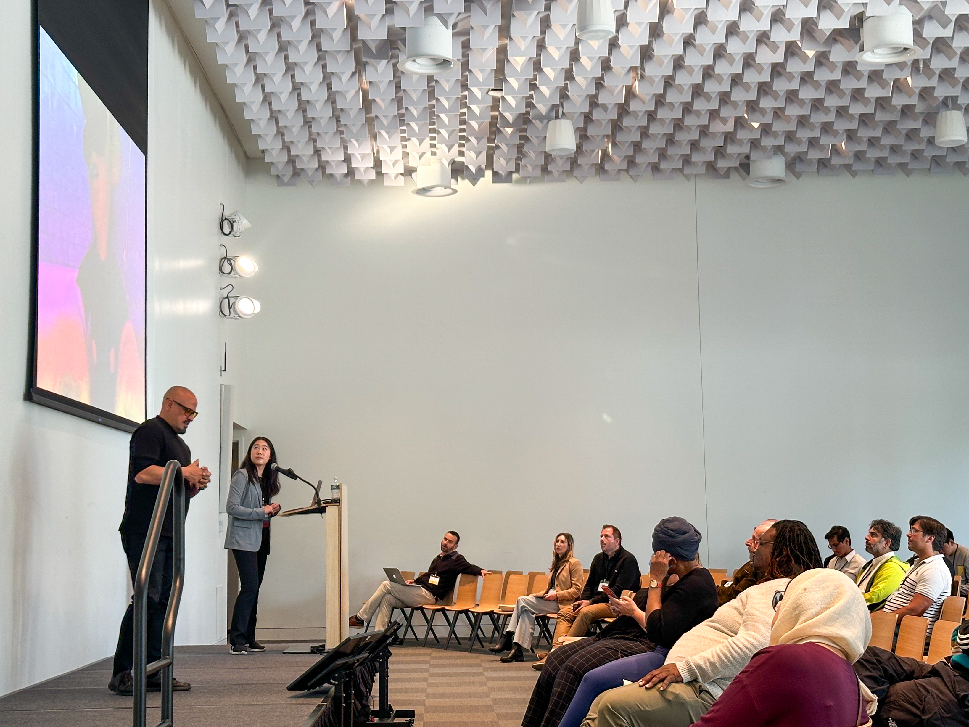 Lucy and an ASL interpreter stand on the stage while audience members watch a video during her paper presentation. The screen shows a still of Luigi from the Mario Brothers franchise.