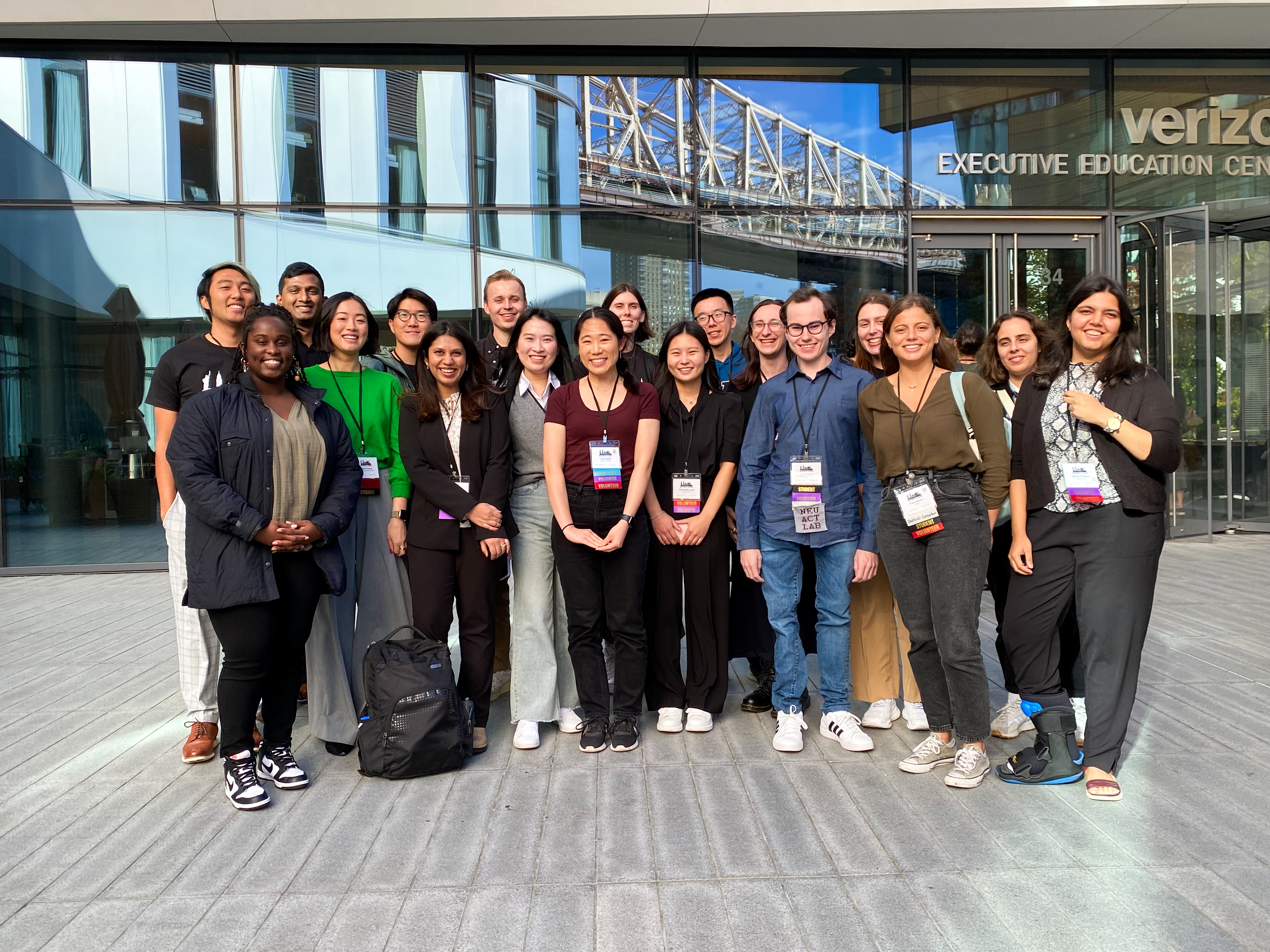 18 of the ASSETS SVs pose for a group photo in front of the conference center, with a reflection of the Queensboro Bridge in the glass windows behind them.