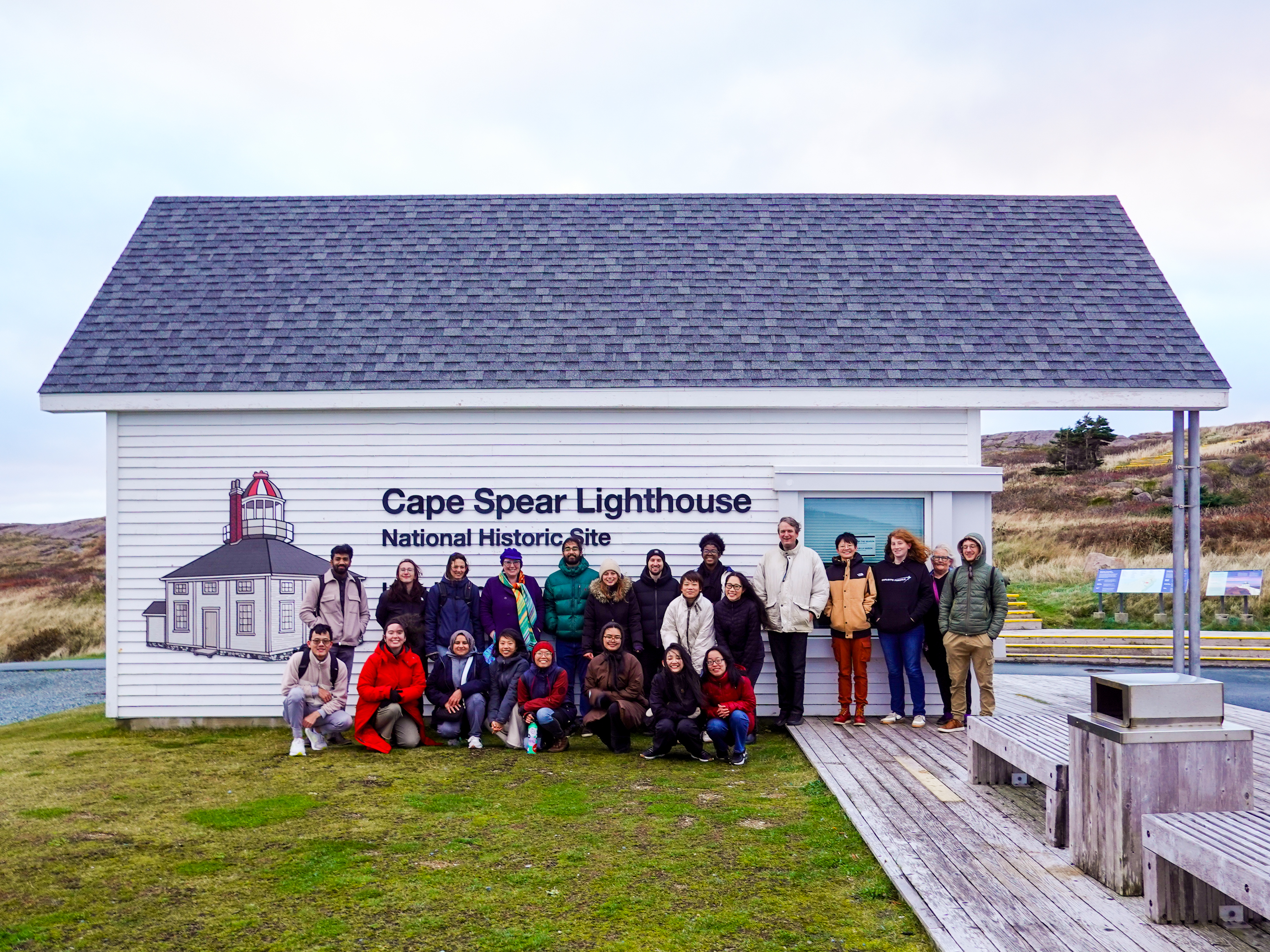 A group of 23 ASSETS attendees wearing colorful winter coats gather in front of a small white building. The side of the building features an illustration of a lighthouse and reads 'Cape Spear Lighthouse National Historic Site'.