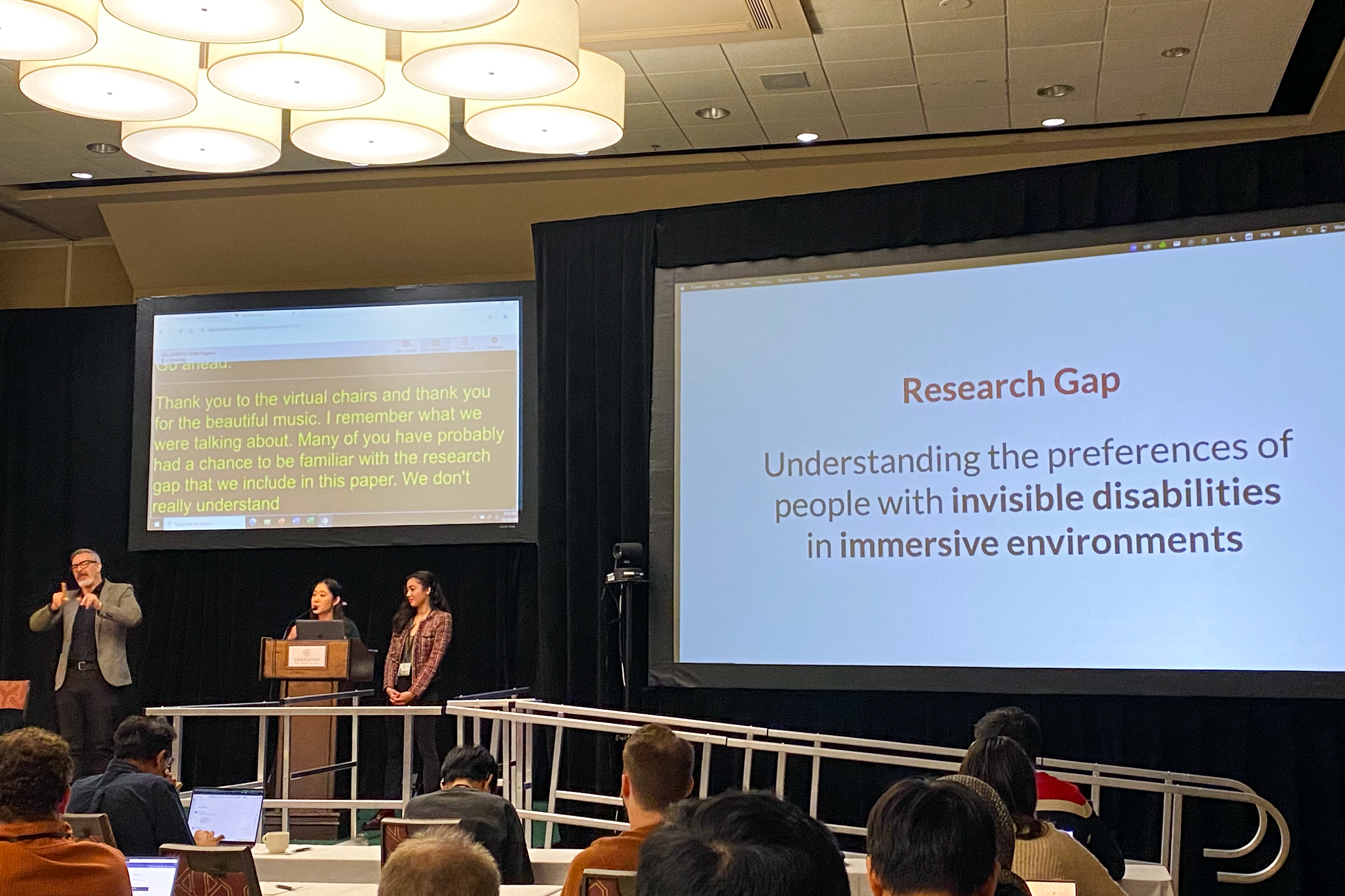 Lucy and Ria stand behind a podium on stage. Lucy is speaking and there is an interpreter to the presenters' right. Behind them is a small projector screen with captions reading 'Thank you to the virtual chairs and thank you for the beautiful music. I remember what we were talking about. Many of you have probably had a chance to be familiar with the research gap that we include in this paper. We don't really understand...'. On the right side of the stage is a larger screen reading 'Research Gap: Understanding the preferences of people with invisible disabilities in immersive environments'.