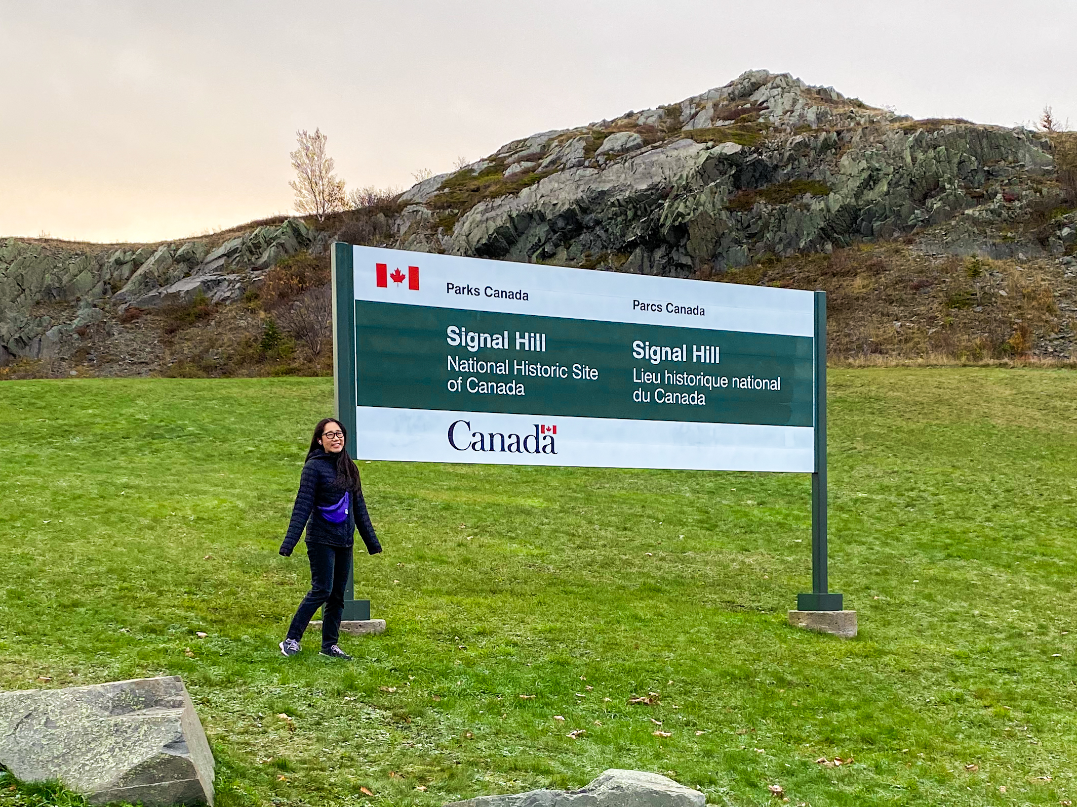 Lucy stands in front of a large white and green sign reading 'Parks Canada, Signal Hill National Historic Site of Canada' in English and French. The sign is planted in a patch of grass and there is a rock formation in the background.