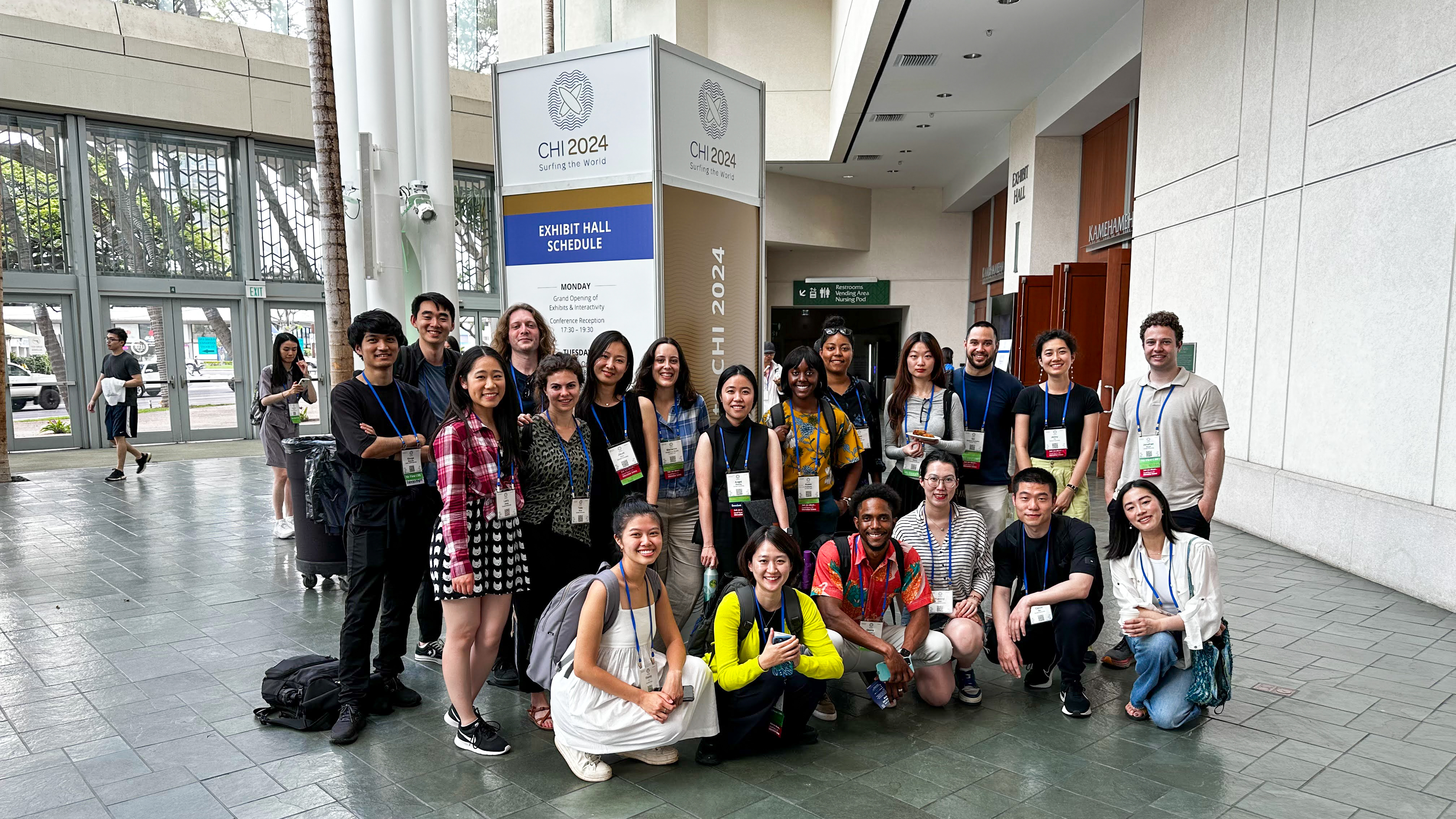 A group photo of 20 Cornell students in the foyer of the Hawai'i Convention Center. Behind them is a tall rectangular display reading 'CHI 2024, Surfing the World.'