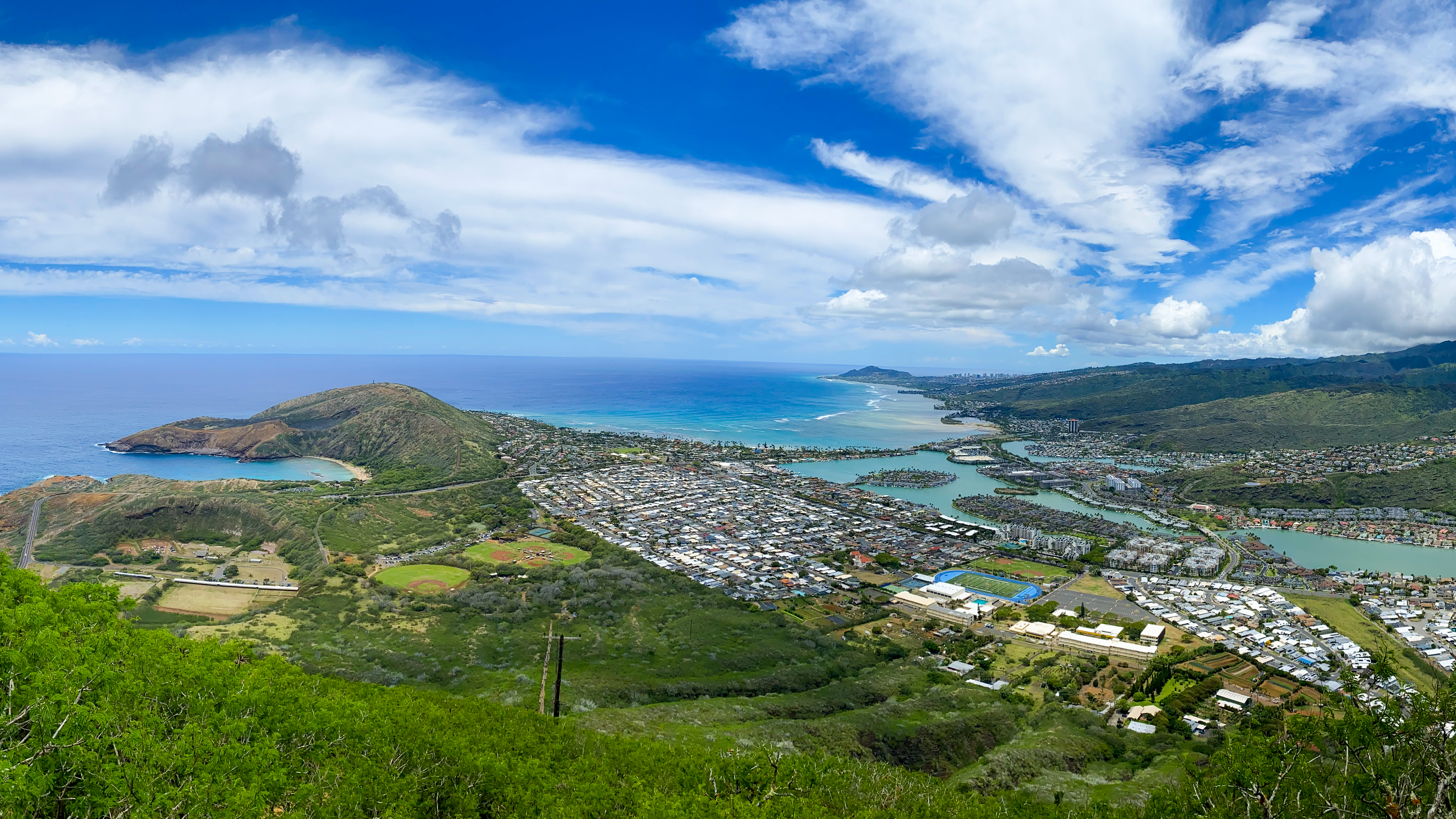 A panoramic photo of the westward view from the summit of the Koko Crater Stairs. There is a small peninsula surrounded by a bright blue ocean on the left, baseball fields and many colorful houses in the center, and dark green mountains and smaller islands surrounded by turquoise water on the right. In the foreground, there is lush green foliage and a thin, light brown line showing the Koko Crater Stairs.