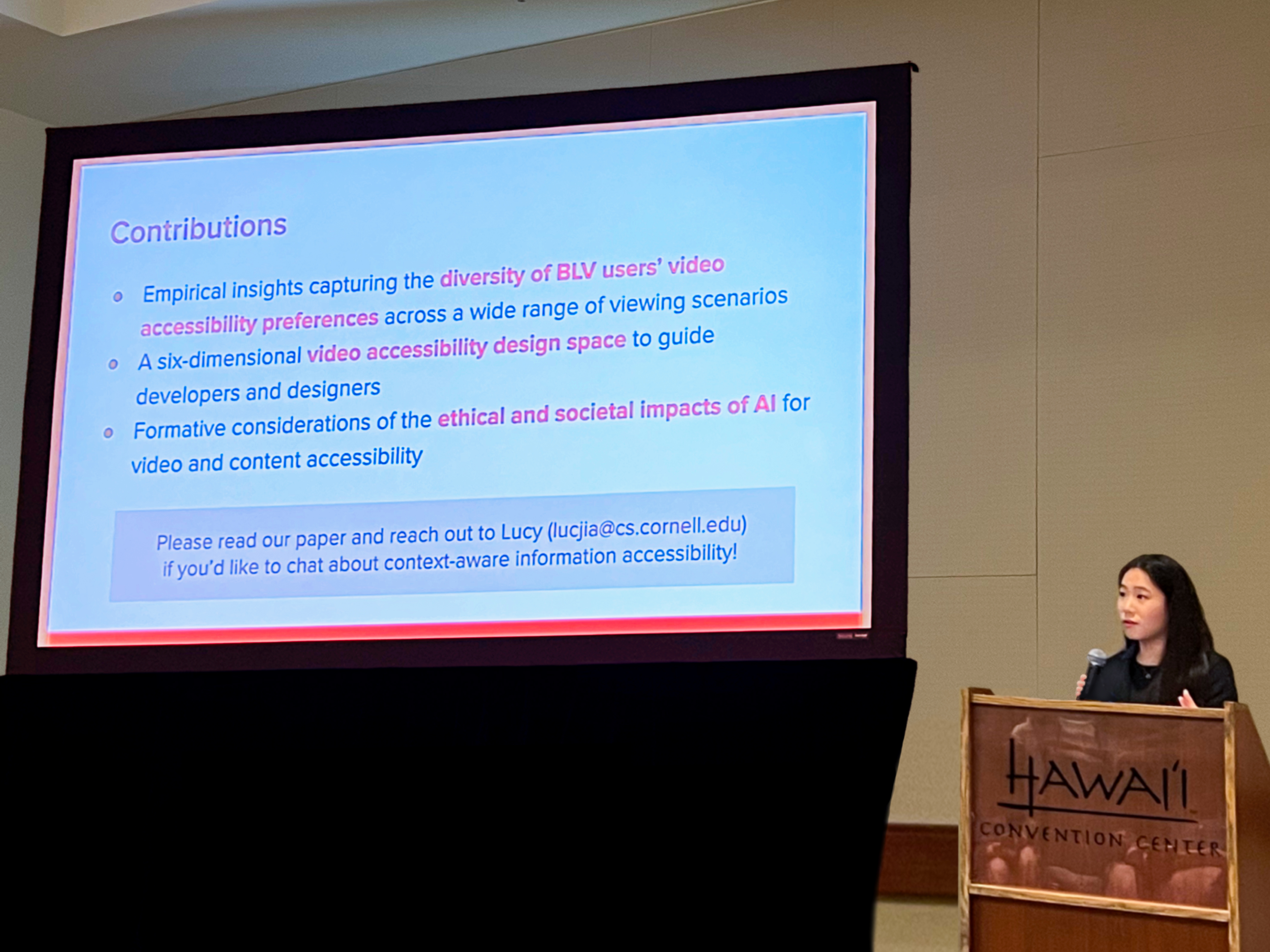 Lucy stands behind a podium saying 'Hawai'i Conference Center.' To her left is a large projector screen showing a Contributions slide, reading: 'Empirical insights capturing the diversity of BLV users' video accessibility preferences across a wide range of viewing scenarios; a six-dimensional video accessibility design space to guide developers and designers; formative considerations of the ethical and societal impacts of AI for video and content accessibility,' encouraging people to read the paper, and containing her contact information.