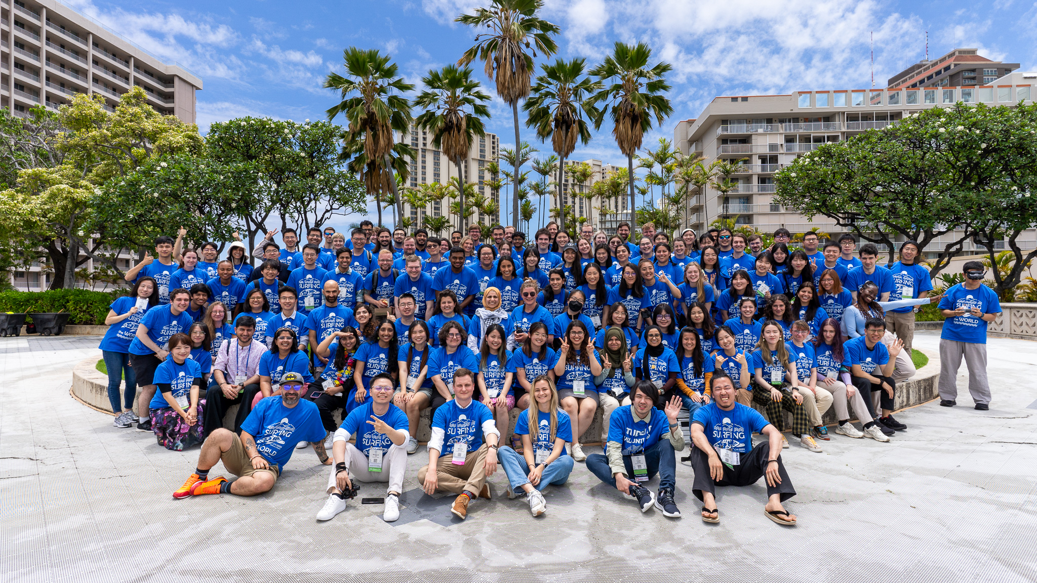 The CHI SVs, a group of approximately 120 people wearing matching blue SV T-shirts, pose for a group photo. There are palm trees and other foliage behind the SVs, and hotels in the distance.