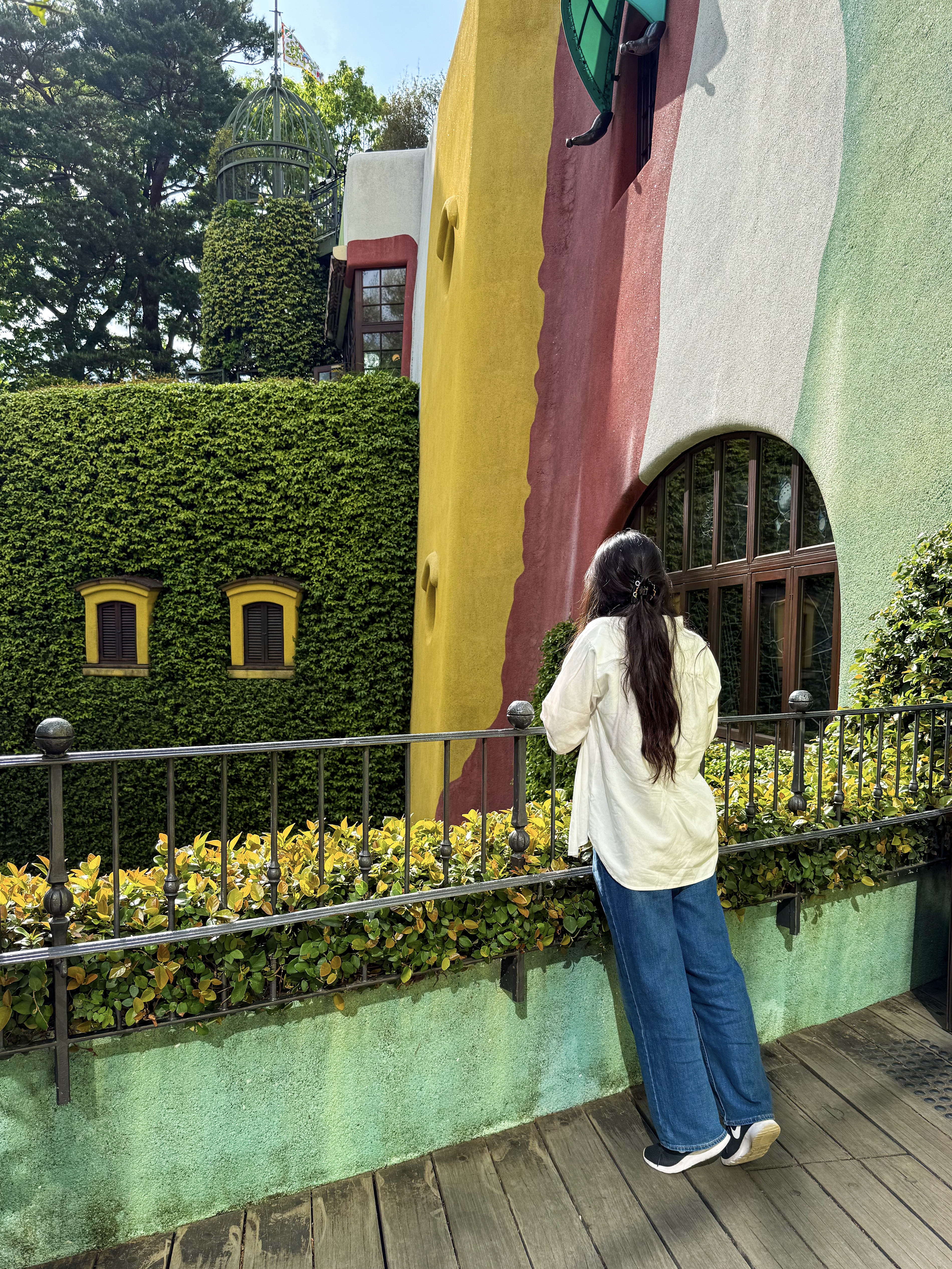 Lucy leans against a black metal railing, faced away from the camera, with multi-colored walls on her right and a wall covered with lush green foliage before her. She is wearing a white linen overshirt, blue jeans, and a soot sprite claw clip in her hair.