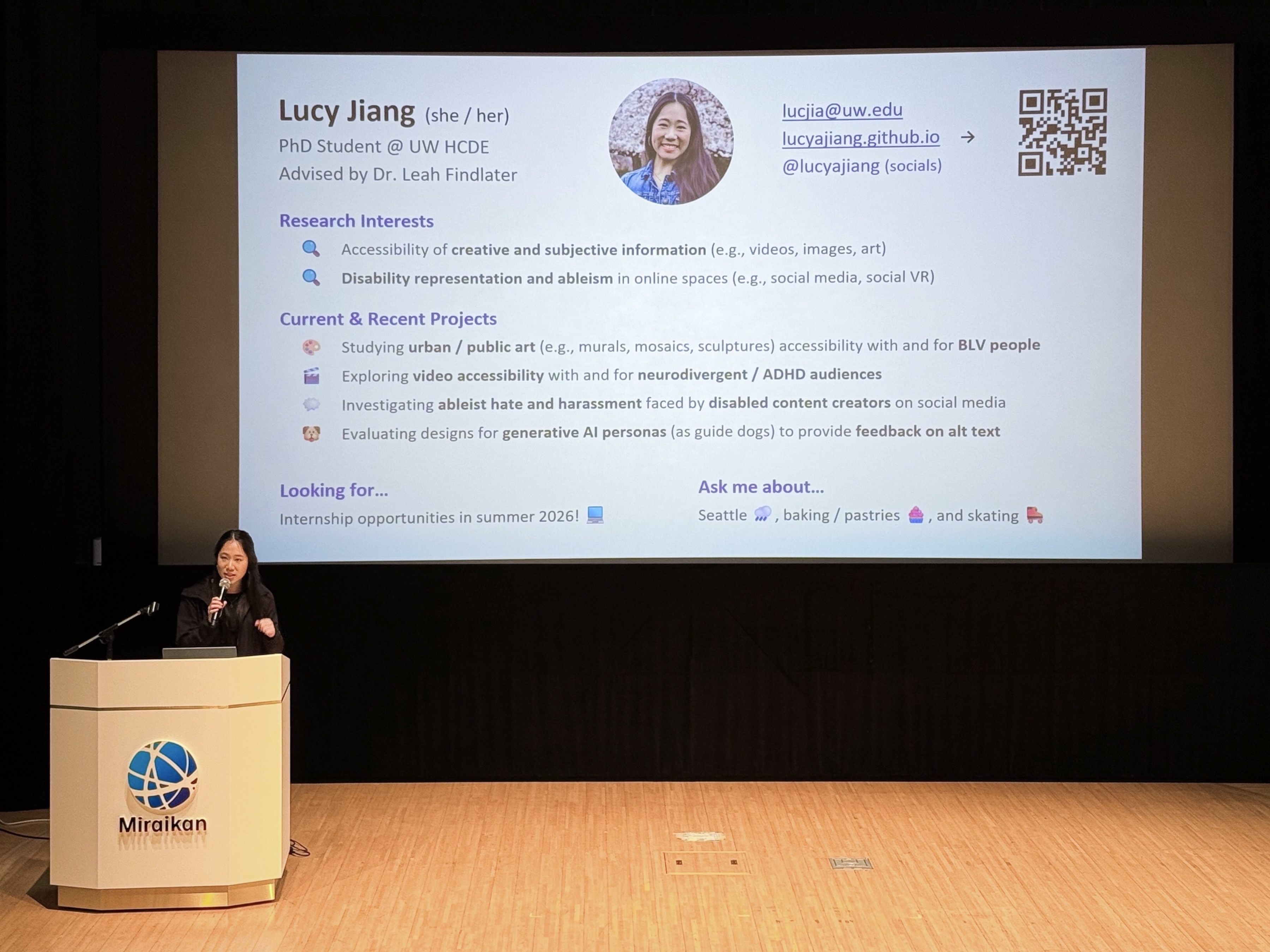 Lucy stands behind a podium with the Miraikan logo, a blue circle with curved lines across it, and presents a lightning talk slide on stage. The slide shows her Research Interests, Current & Recent Projects, and her contact information.