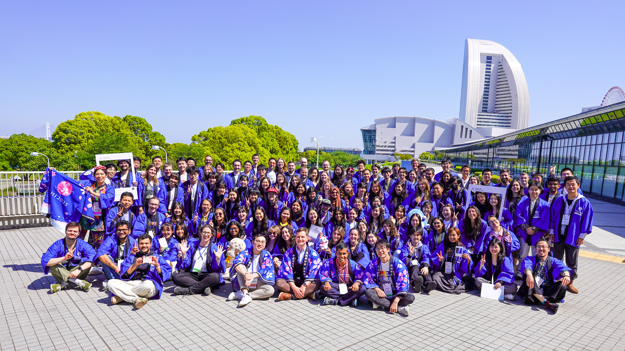 The CHI SVs, a group of approximately 120 people wearing matching blue SV happis (a traditional Japanese jacket), pose for a group photo. Some SVs hold up large cardboard frames resembling the Instagram interface. Behind the SVs is the Minato Mirai skyline, composed of white and grey geometric buildings and bright green trees.