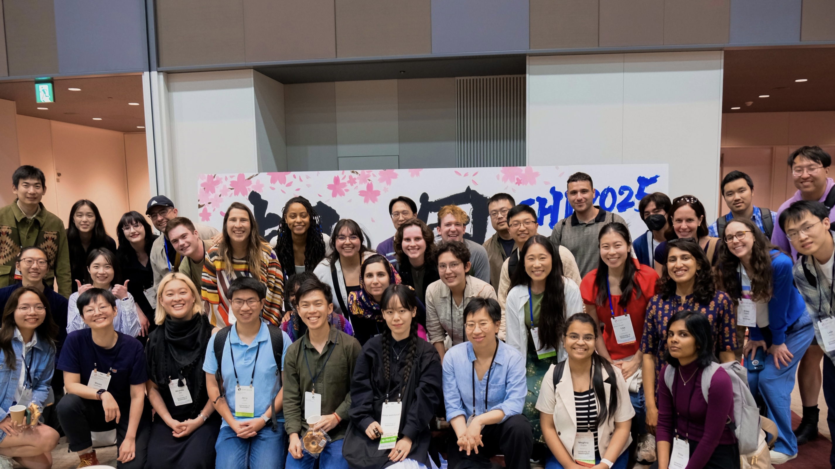 A group photo of 36 UW students, faculty, and graduates at the convention center. Behind them, but mostly obscured by the group, is a large rectangular display with cherry blossom illustrations and the words 'CHI 2025' in blue calligraphy.