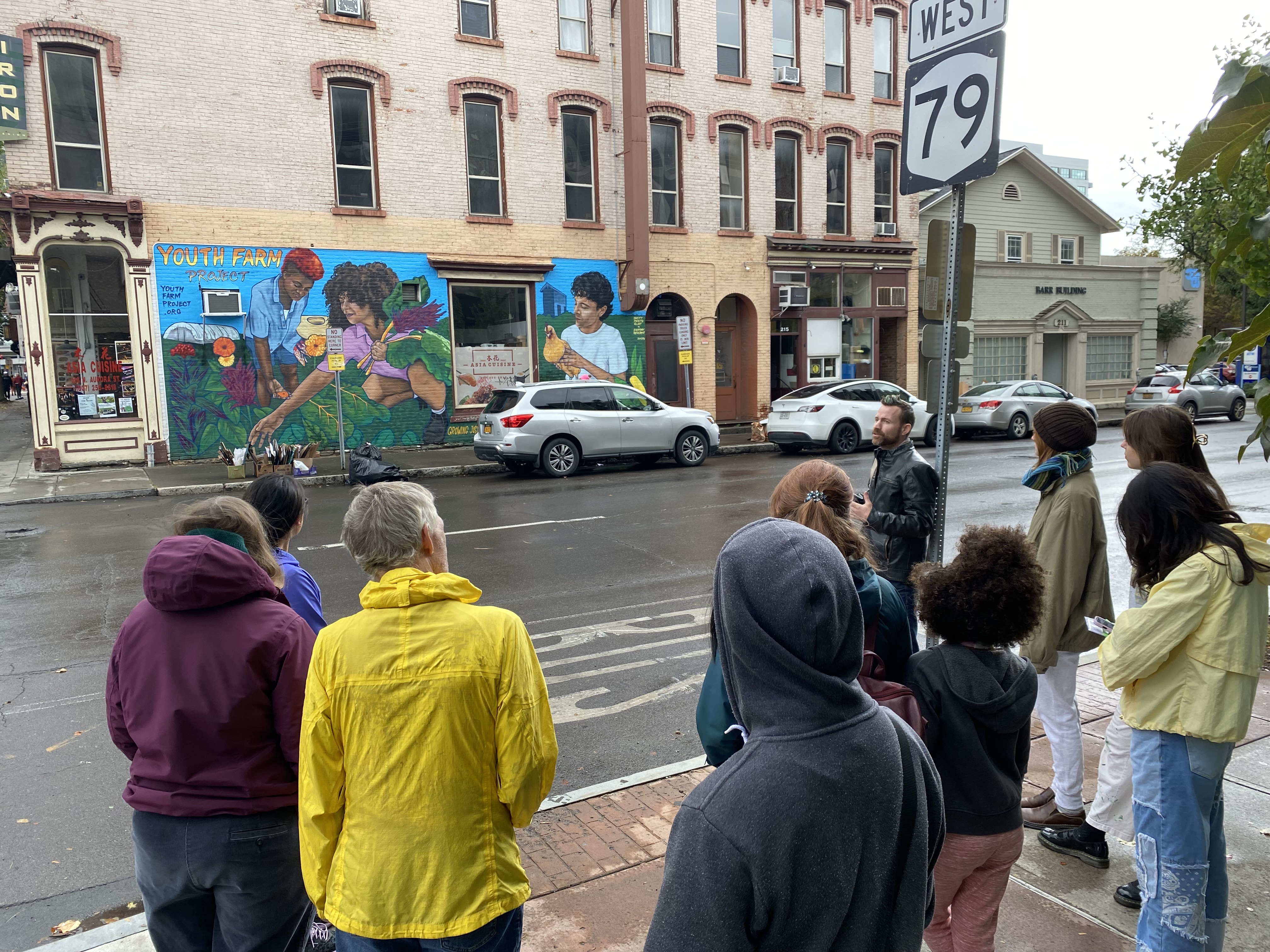 Ten mural tour attendees stand with their backs to the camera, across the street from the 'Youth Farm Project' mural. The mural is a large piece approximately one story tall and two car-lengths wide, and was painted by Efren Rebugio and Brittany Johnson. At the top left, orange and yellow text reads 'Youth Farm Project'. From left to right, there are three people of color farming: a woman with an orange and red fro-hawk collects wildflowers, a woman with a fluffy afro gathers chard, and a man holds a chicken in his hands. Caleb, a white person with short hair wearing a leather jacket, speaks in front of the group.