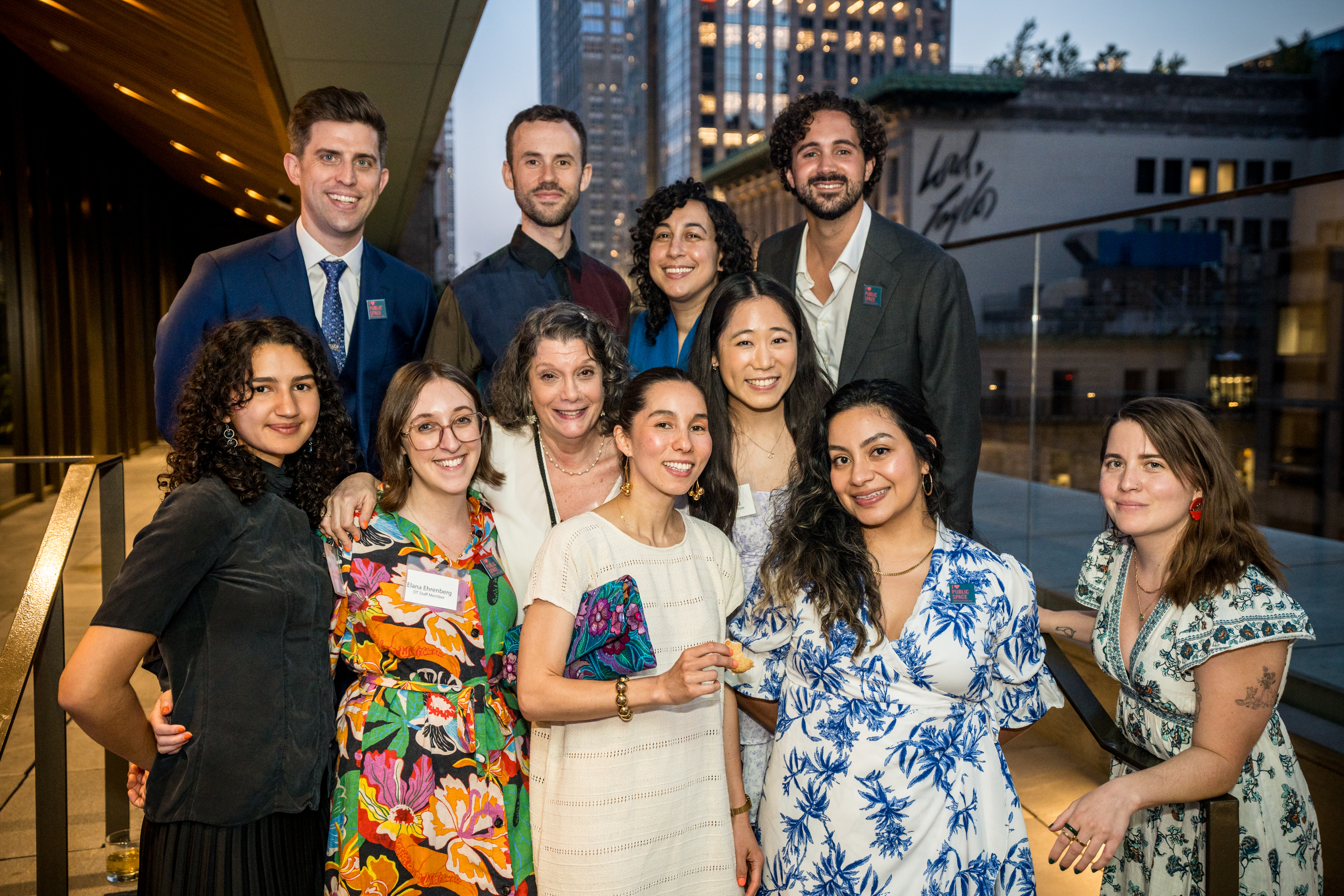 Group photo of 11 staff, fellows, and interns from the Design Trust for Public Space at the annual Design Trust Benefit, held at the Stavros Niarchos Foundation Library rooftop. The group members are wearing semi-formal attire such as suits and floral dresses, and all have wide smiles. In the background, there is a building with a sign reading 'Lord & Taylor' and a glass skyscraper behind it.