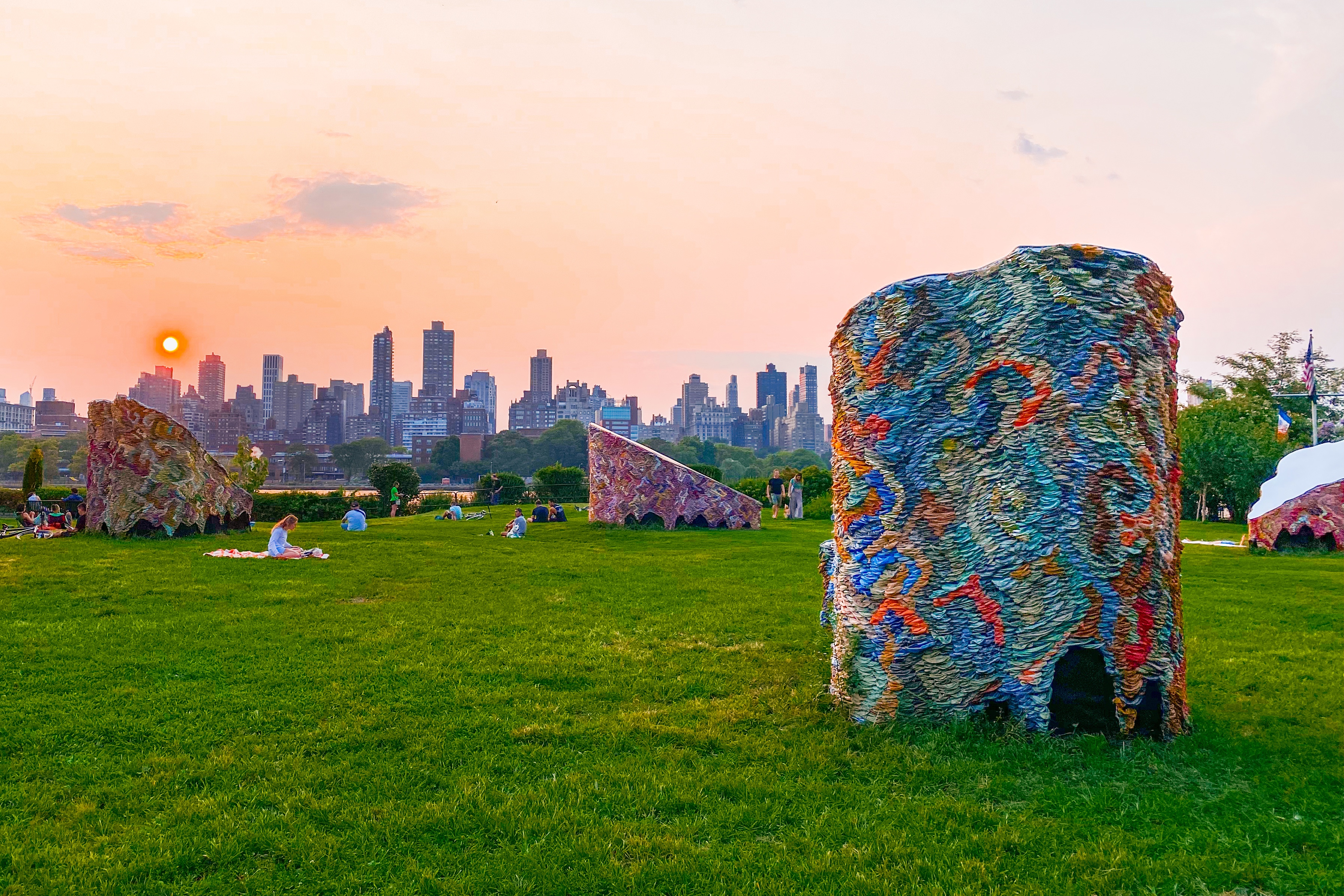 Socrates Sculpture Park at sunset, with a large sculptural art piece in the foreground, three similar sculptural pieces and people sitting on grass and walking around the park in the midground, and an orange sunset over the Manhattan skyline in the background. The sculptural pieces, titled 'We are Nomads, We are Dreams' and created by Suchitra Mattai, are draped with colorful saris woven together. This park was the site of Design Trust's August 2024 Public Space Potluck.